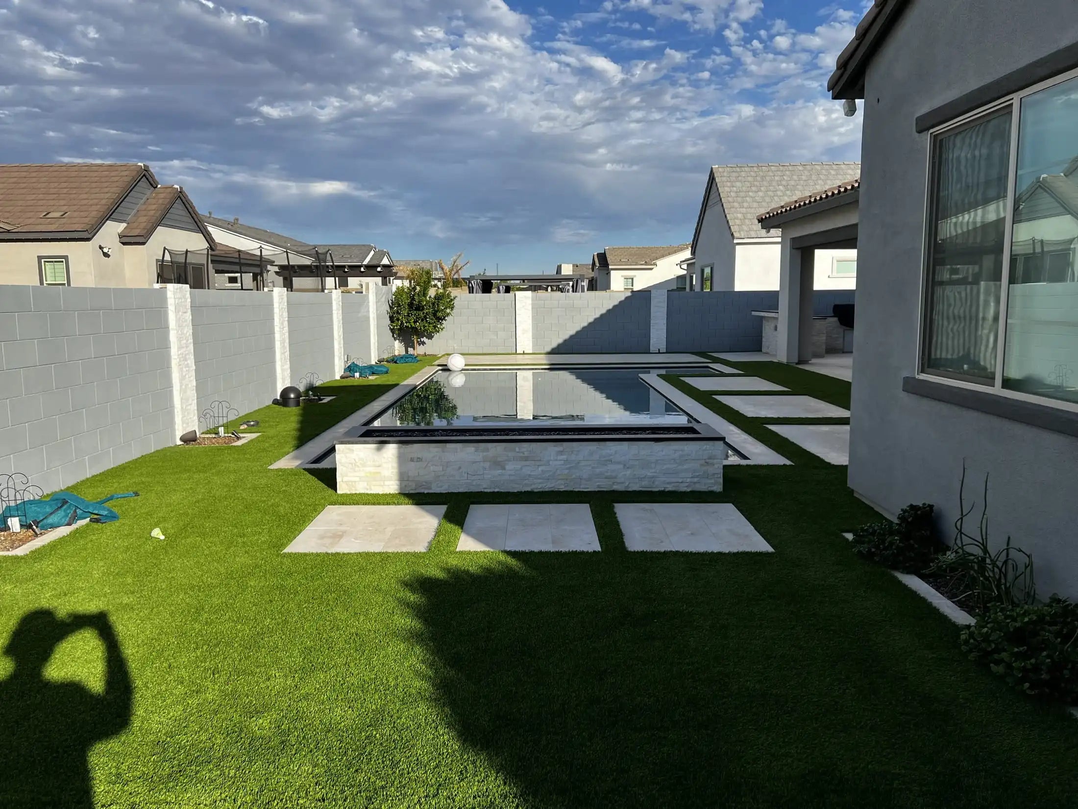 Backyard with a pool, grass, and neighboring houses under a blue sky.